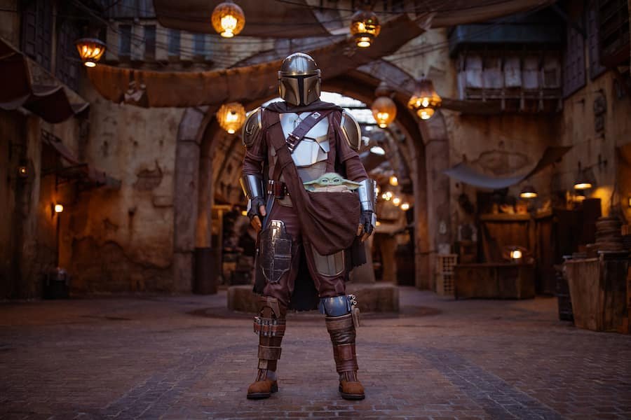 Mandaloriano de armadura e capacete carregando Grogu em cenário temático de Star Wars.
